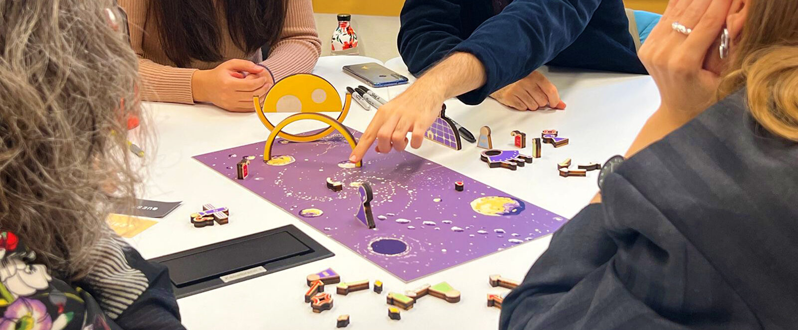 Four people playing UNBOX on the space board, using metaphorical pieces such as an alien, rings, and fuel.